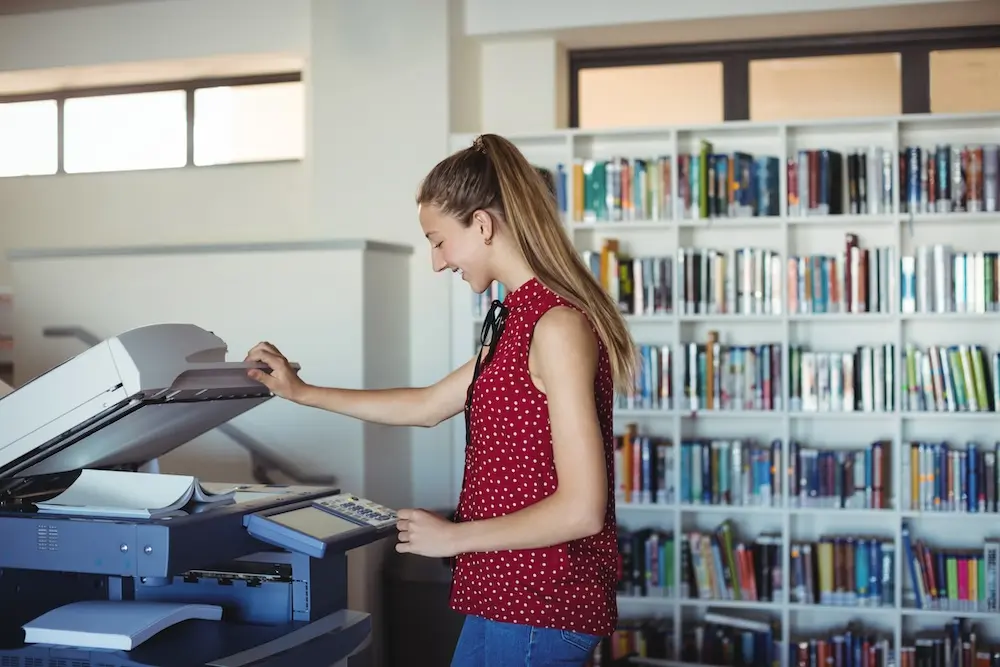 Automated energy data management - A young woman wearing a red sleeveless polka dot blouse uses a photocopier in a library. Bookshelves filled with books are visible in the background.