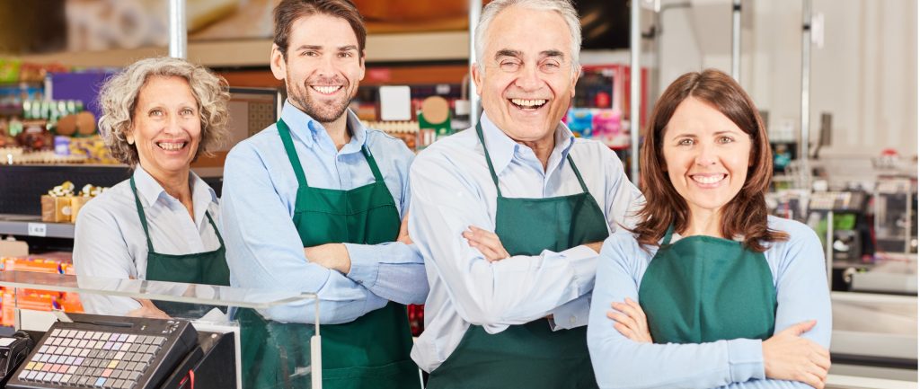 Automated energy management - Four supermarket employees wearing green aprons and light blue shirts stand side by side with arms folded, smiling at the camera. A till and shop shelves are visible in the background. Automated energy management - Four supermarket employees wearing green aprons and light blue shirts stand side by side with arms folded, smiling at the camera. A till and shop shelves are visible in the background.