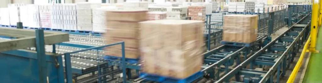 Automated energy management - Conveyor system transporting pallets wrapped in plastic through a warehouse or distribution centre, with multiple packages being moved along parallel conveyor belts.