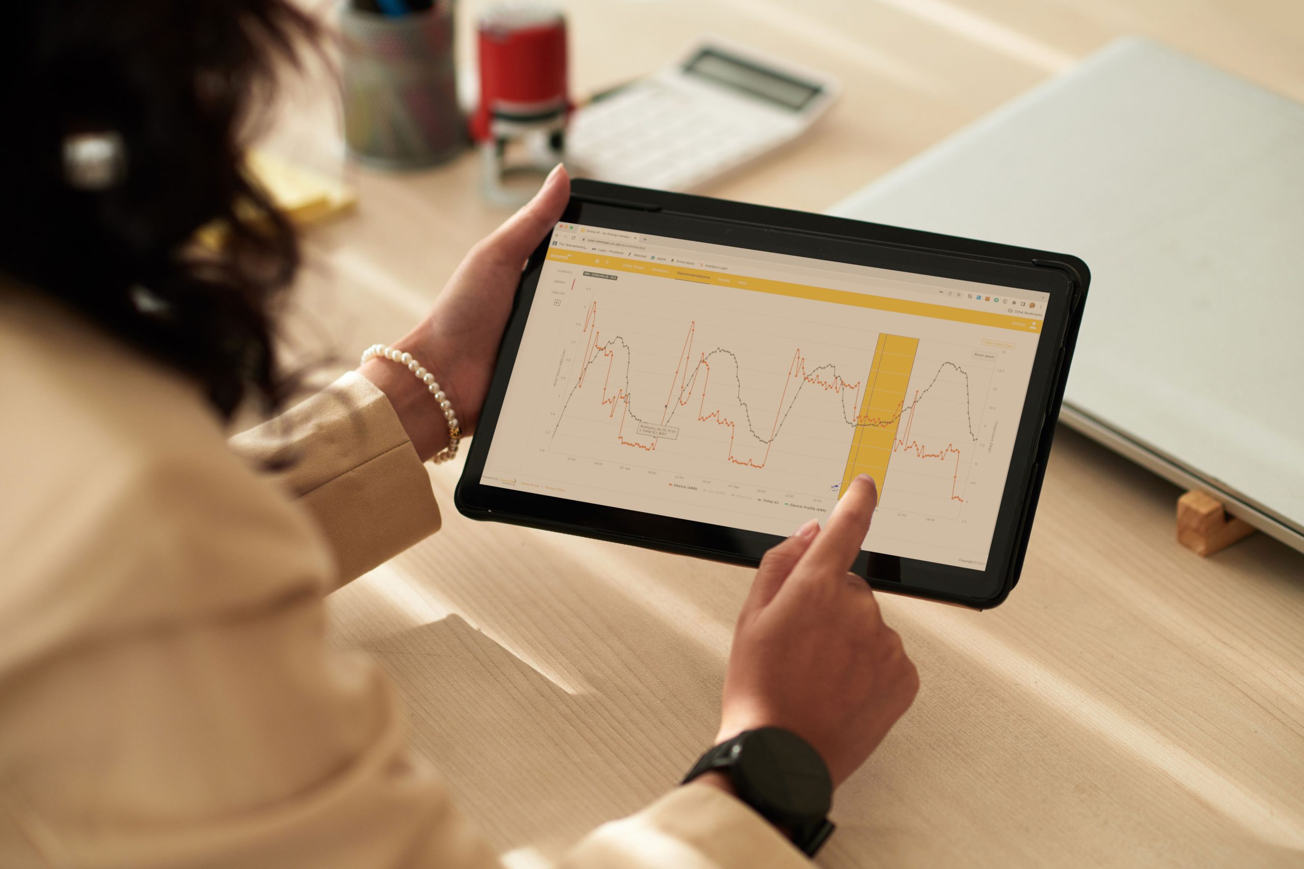 Automated energy management - A person holds a tablet displaying a line graph with fluctuating data trends and highlights a section in yellow. The scene includes a calculator, notebook, and office supplies on a light desk.