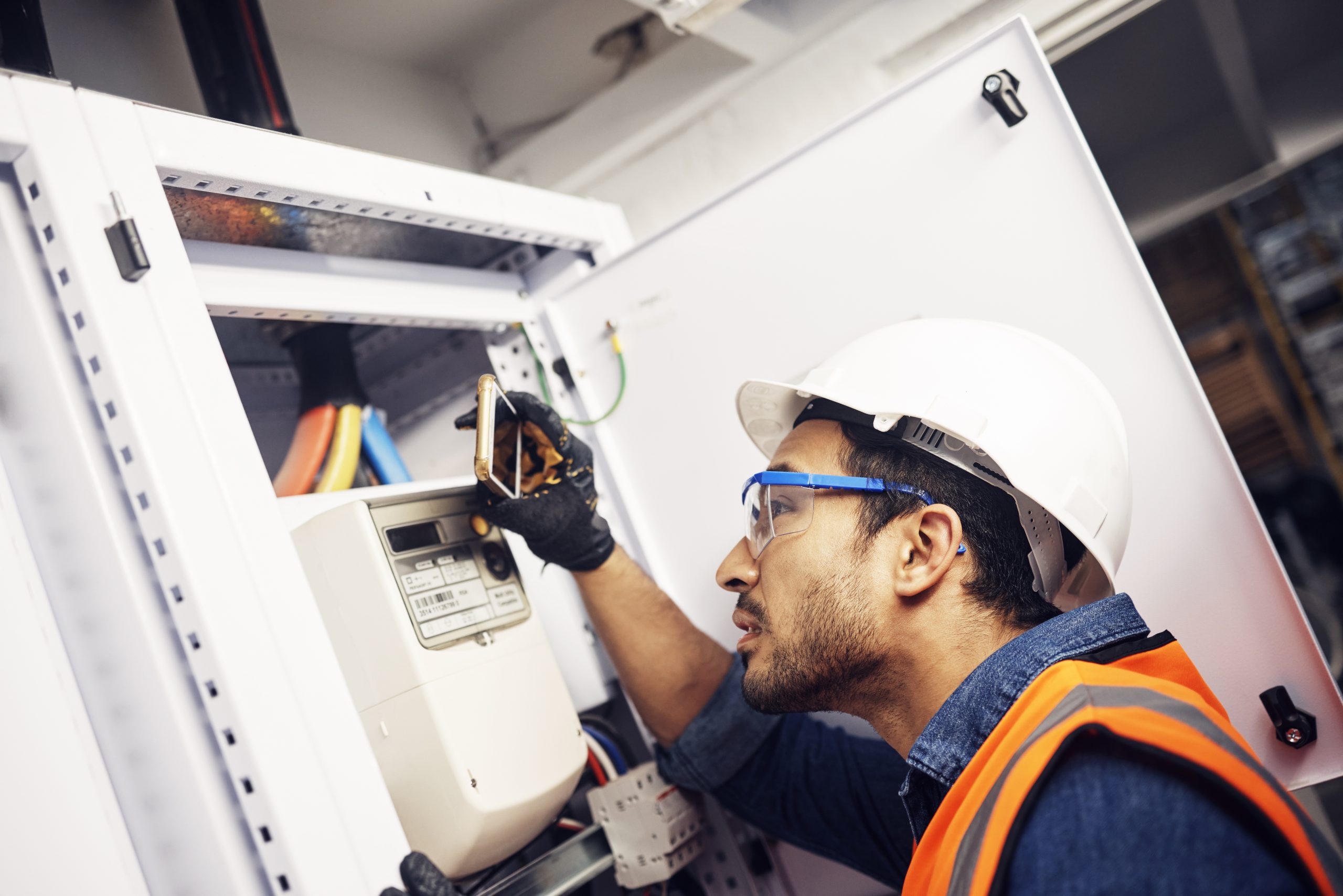 Automated energy management - A man wearing a white hard hat, safety glasses, and an orange safety vest examines an electricity meter inside a control panel, holding a tool and inspecting the wiring.