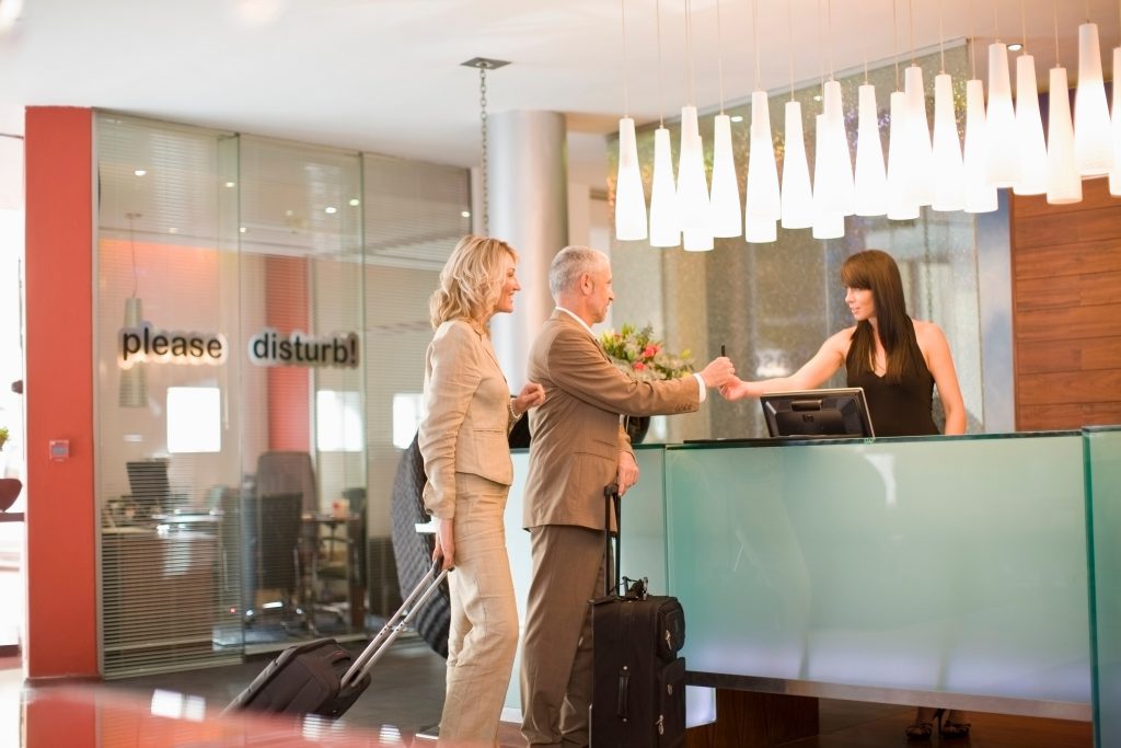 Automated energy management - hotel energy saving - A man and a woman with suitcases stand at a hotel reception desk while a receptionist hands the man a key card. The lobby is modern, with hanging lights and a glass wall bearing the words please disturb.