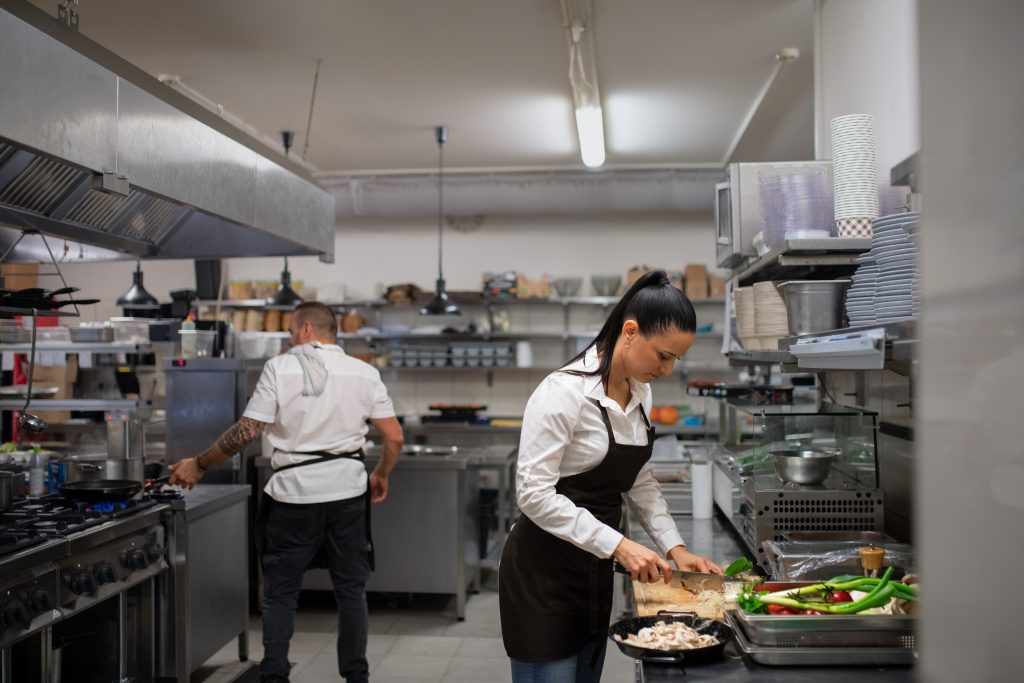 Automated energy management - restaurant hospitality energy saving - A woman in an apron chops vegetables at a counter in a commercial kitchen, while a man in an apron works at a cooker in the background. The kitchen is organised with shelves, crockery, and cooking equipment visible.