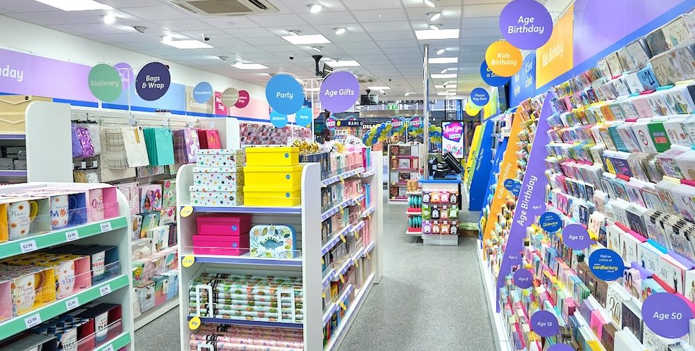 Automated energy management - A brightly lit shop aisle displaying colourful cards and gift wrap on shelves, with hanging signs categorising items such as “Party,” “Age Birthday,” and “Gift Bags and Wrap.” The floor is grey and the ceiling has white tiles.