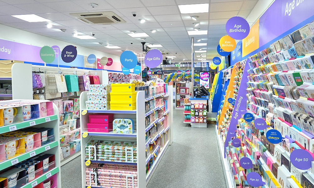 Automated energy management - A brightly lit shop aisle displaying colourful cards and gift wrap on shelves, with hanging signs categorising items such as “Party,” “Age Birthday,” and “Gift Bags and Wrap.” The floor is grey and the ceiling has white tiles.