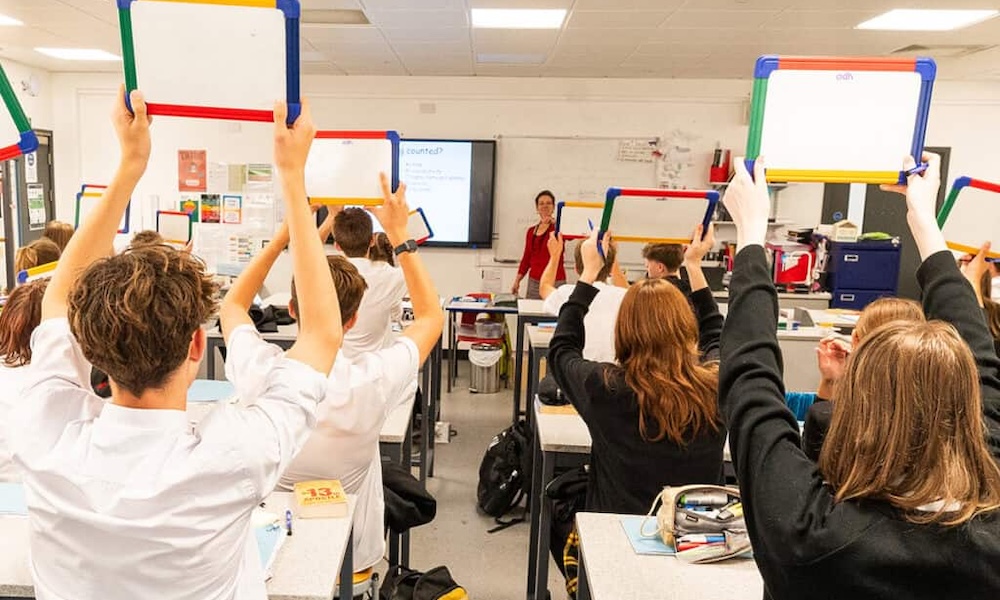 Automated energy management - Students in a classroom sit at desks, holding up small whiteboards. A teacher stands at the front near a projector screen, addressing the class. The room is brightly lit with various supplies and posters visible.