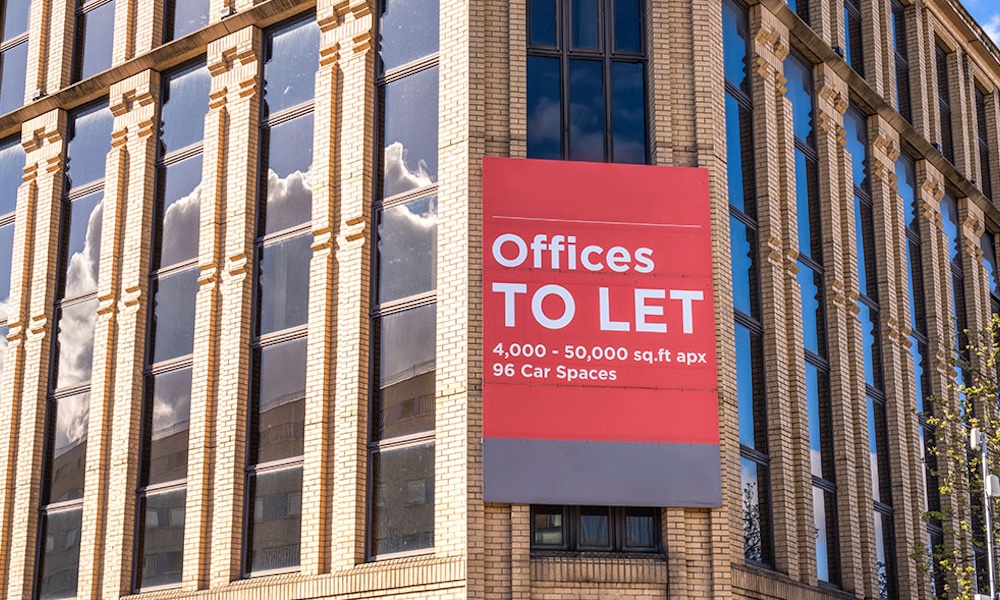 Automated energy management - A brick office building with tall windows and a large red sign that reads, “Offices TO LET, 4,000 – 50,000 sq. ft. approx., 96 Car Parking Spaces.” The sign is mounted on the corner of the building.