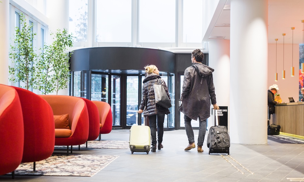 Automated energy management - Two people with suitcases walk through a modern hotel lobby with red chairs, large windows, and a receptionist at the front desk.