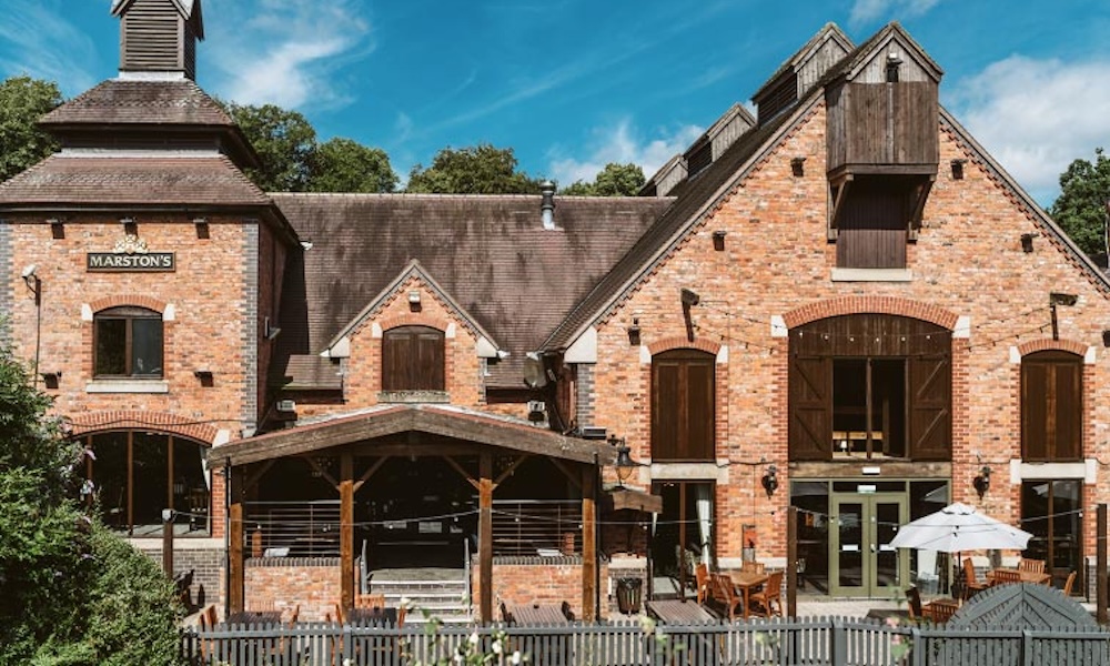 Automated energy management - A large red-brick building with a covered outdoor seating area, wooden balcony, and several windows and doors. The sky is clear and blue, and there are tables and chairs on a patio in front.