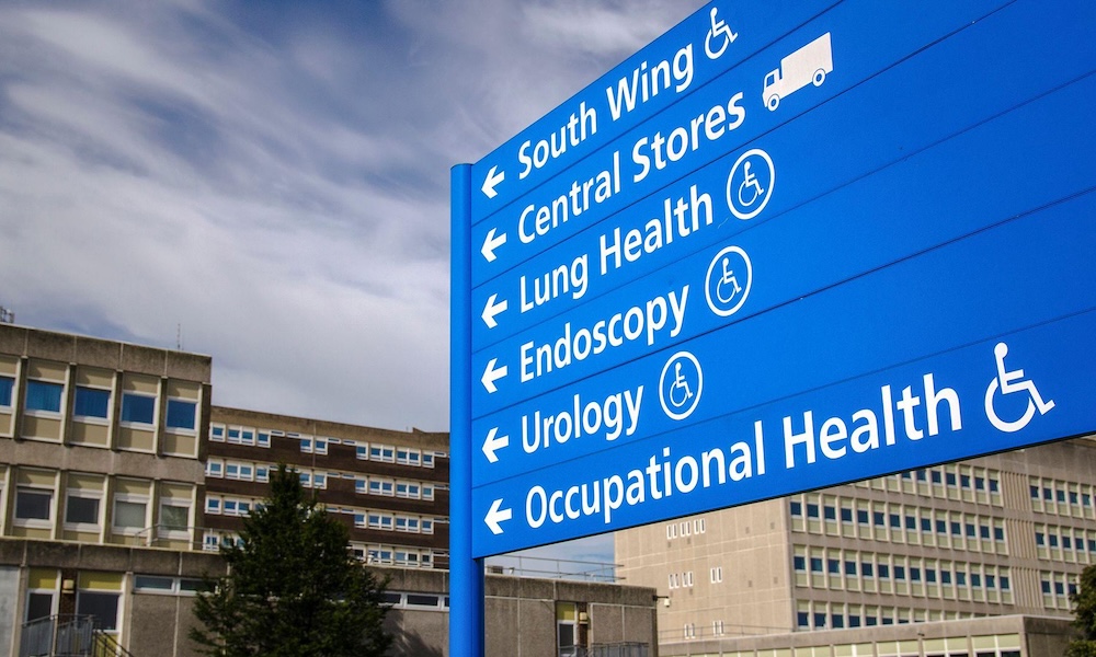 Automated energy management - A blue hospital sign outdoors lists directions to South Wing, Central Stores, Lung Health, Endoscopy, Urology, and Occupational Health, with symbols for accessibility. Hospital buildings are in the background.