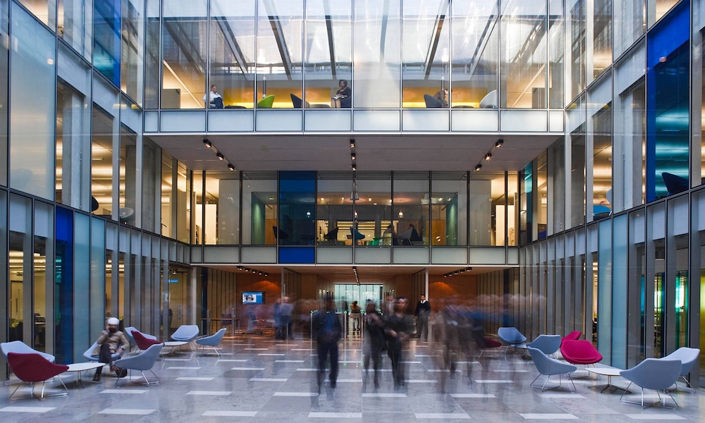 Automated energy management - A modern office atrium with glass walls, several floors, and open seating. People are walking and sitting in the reception area with colourful chairs, while others work in offices above. The space is bright with natural light.