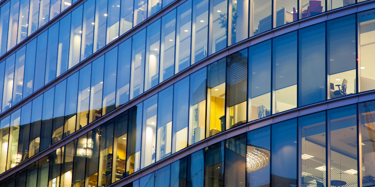 Automated energy management - Close-up of a modern glass office building with curved lines, showing several floors and illuminated rooms inside. Office furniture and lights are visible through the large windows.