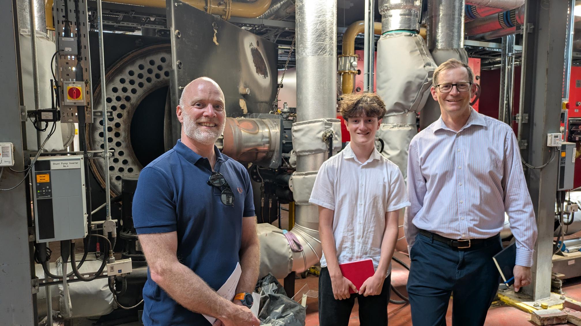 Automated energy management - Three people stand in front of industrial machinery and pipes. One wears a blue polo shirt, another a white short-sleeved shirt, and the third a light dress shirt. Two hold notebooks and all are smiling at the camera. Automated energy management - Three people stand in front of industrial machinery and pipes. One wears a blue polo shirt, another a white short-sleeved shirt, and the third a light dress shirt. Two hold notebooks and all are smiling at the camera.